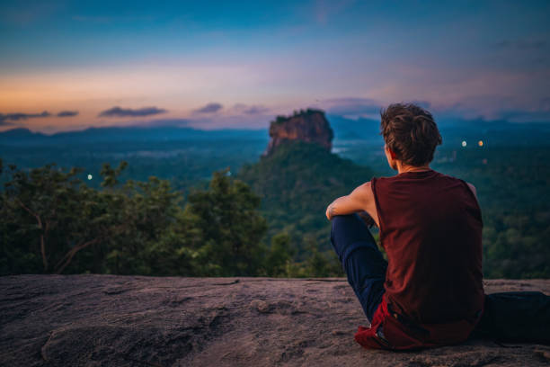 Rear view of a young man sitting on top of a rock at sunrise and contemplating